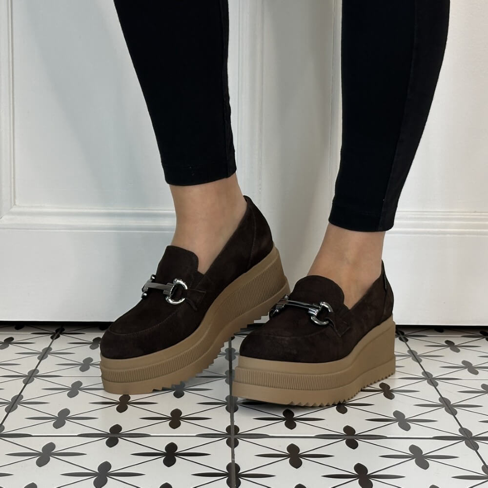 A person wearing Marco Moreo dark brown platform slip-on shoes with a small silver horse bit detail on the strap, paired with black leggings, standing on a black and white patterned tile floor.