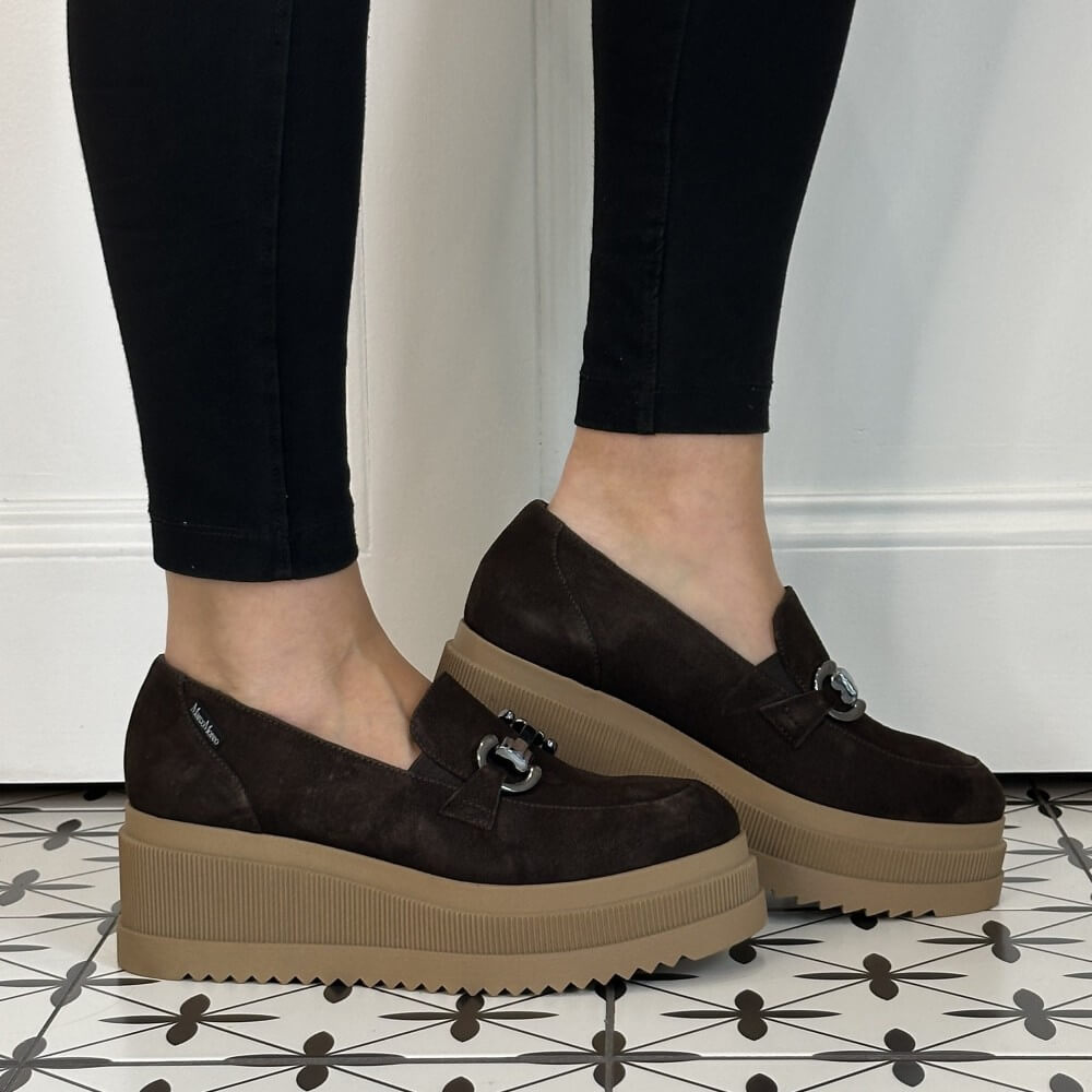 A person wearing Marco Moreo dark brown platform slip-on shoes with a small silver horse bit detail on the strap, paired with black leggings, standing on a black and white patterned tile floor.