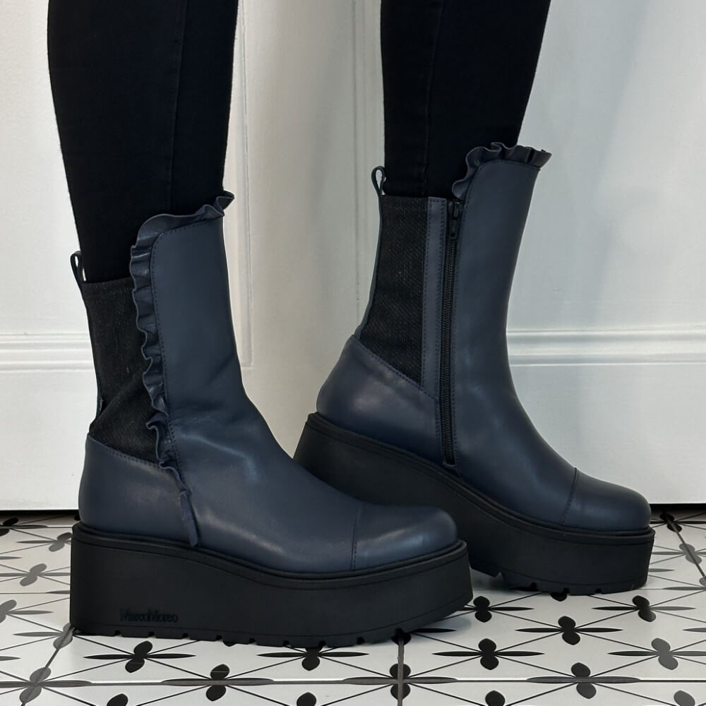 A pair of Marco Moreo dark navy blue platform ankle boots with ruffled trim and side zippers, worn with black leggings on a patterned black and white tile floor.