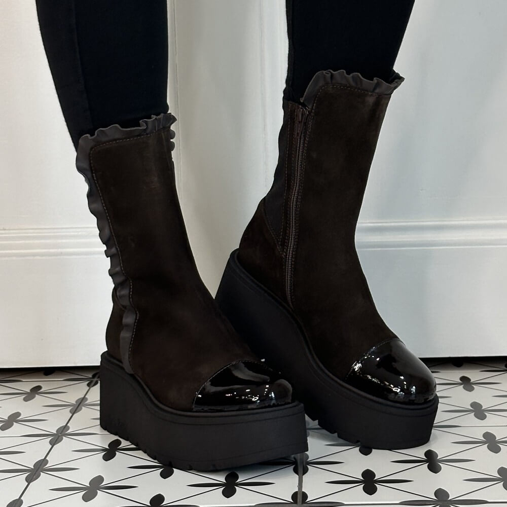 Marco Moreo Dark brown platform ankle boots with a ruffled elastic side panel and glossy toe caps, shown on a person wearing black leggings against a white wall and patterned tile floor.