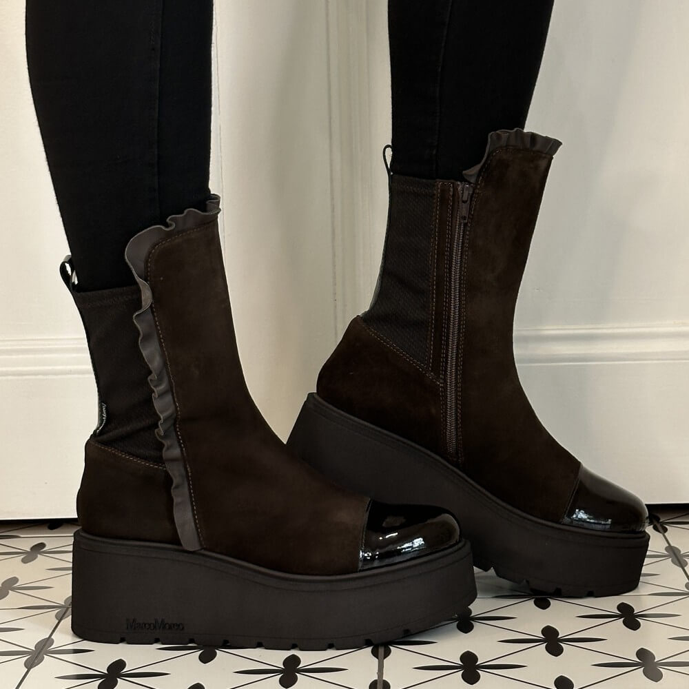 Marco Moreo Dark brown platform ankle boots with a ruffled elastic side panel and glossy toe caps, shown on a person wearing black leggings against a white wall and patterned tile floor.