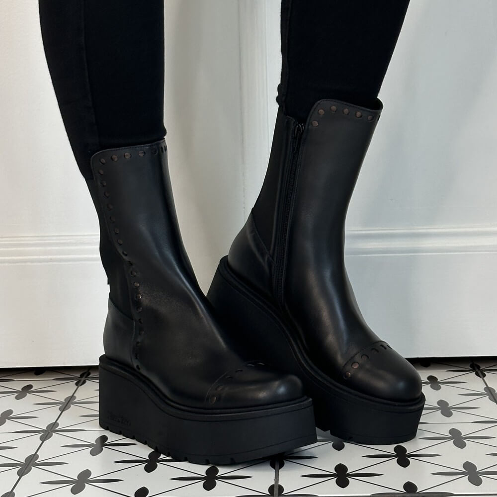 Marco Moreo Black platform ankle boots with elastic side panels and decorative Punched dots, shown worn with black leggings on a patterned tile floor against a white wall.