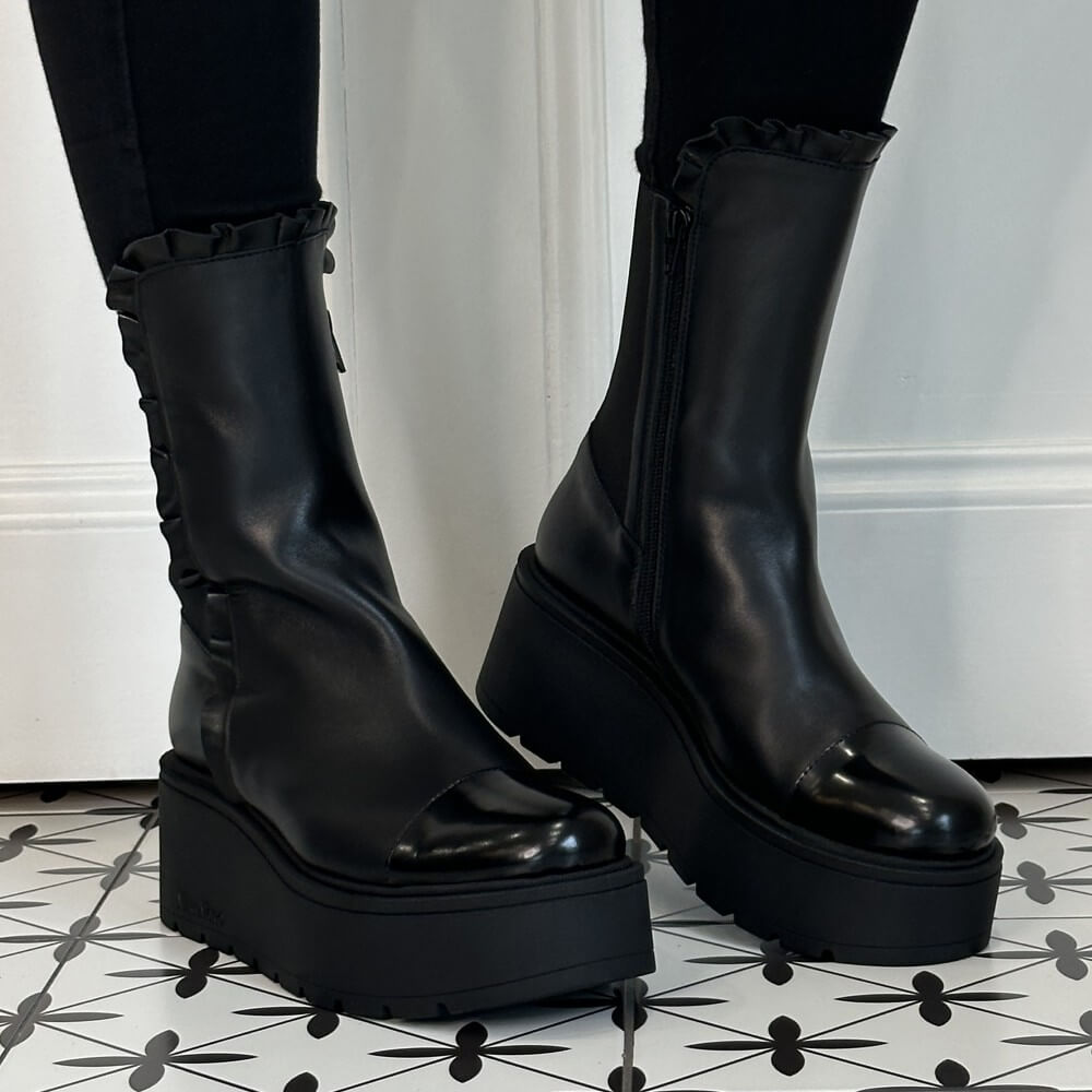 A pair of Marco Moreo black platform ankle boots with ruffled trim at the top, worn with black leggings on a black-and-white patterned tile floor.
