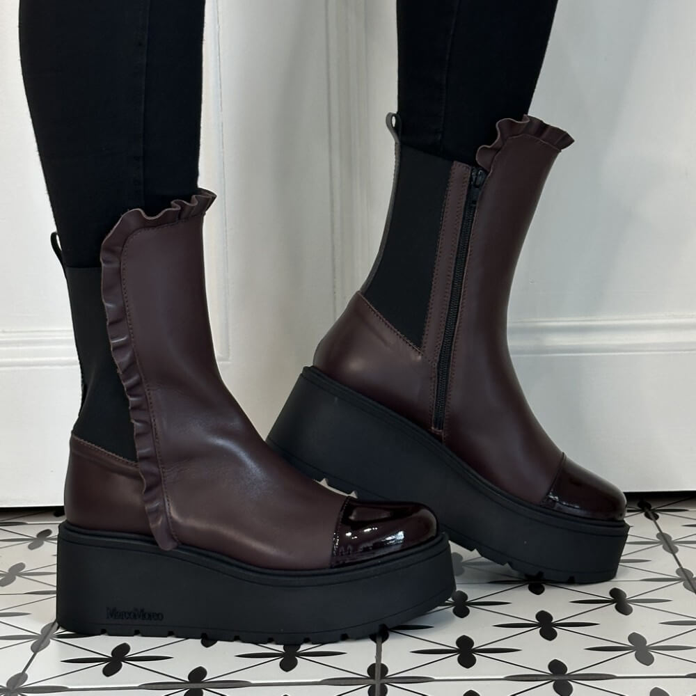 A pair of Marco Moreo dark wine ankle boots with black platform soles, ruffled trim, and side zippers, shown on a model wearing black leggings on a black and white patterned tile floor.