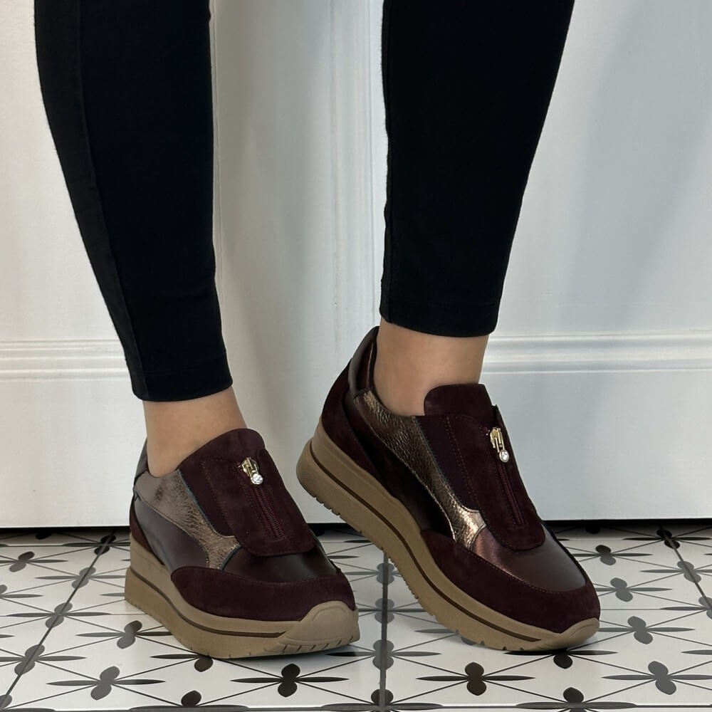 A person wearing Marco Moreo dark burgundy slip-on platform sneakers with metallic silver panels and black leggings, standing on a black-and-white patterned tile floor.