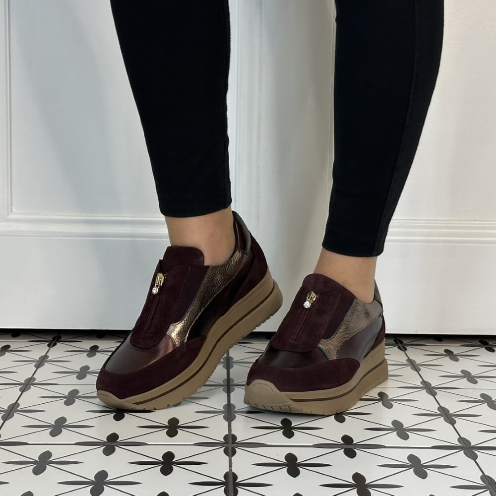 A person wearing Marco Moreo dark burgundy slip-on platform sneakers with metallic silver panels and black leggings, standing on a black-and-white patterned tile floor.