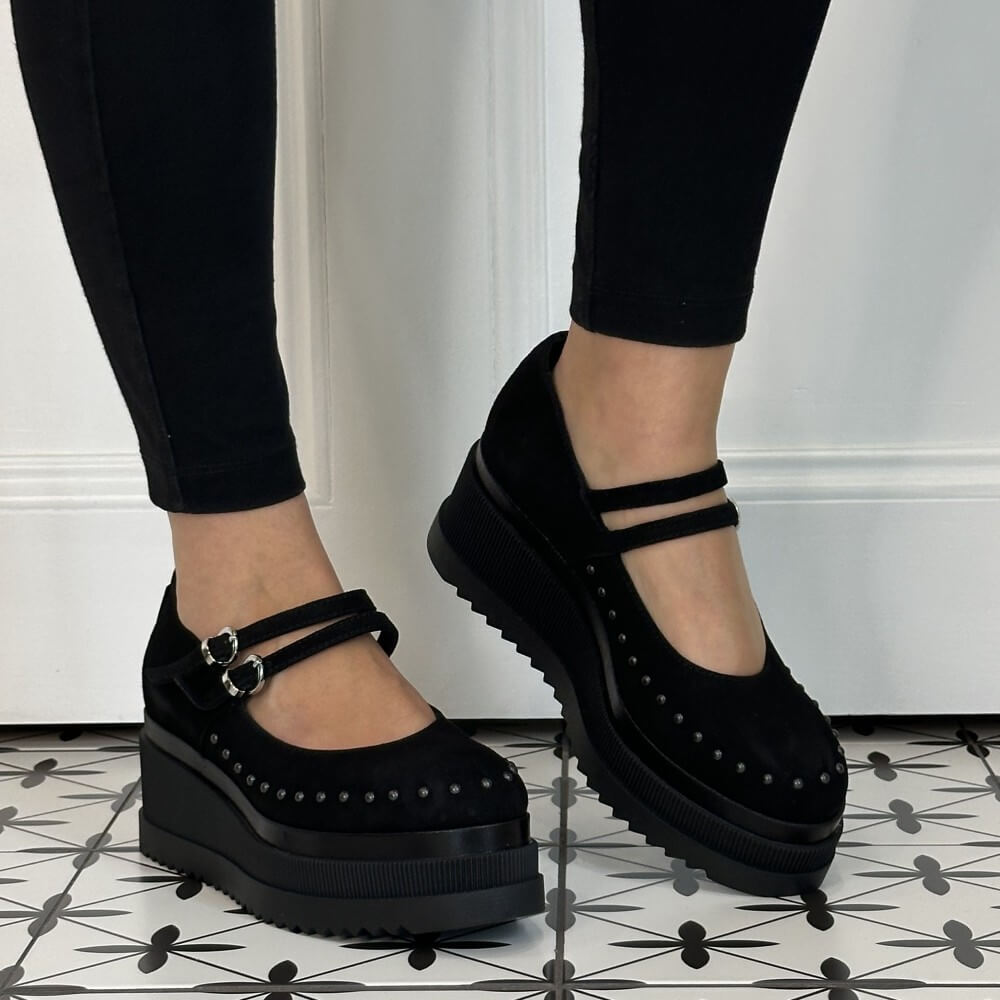 Marco Moreo Black platform Mary Jane shoes with double straps and silver buckles, shown on a model wearing black leggings against a white panelled wall and patterned tile floor.
