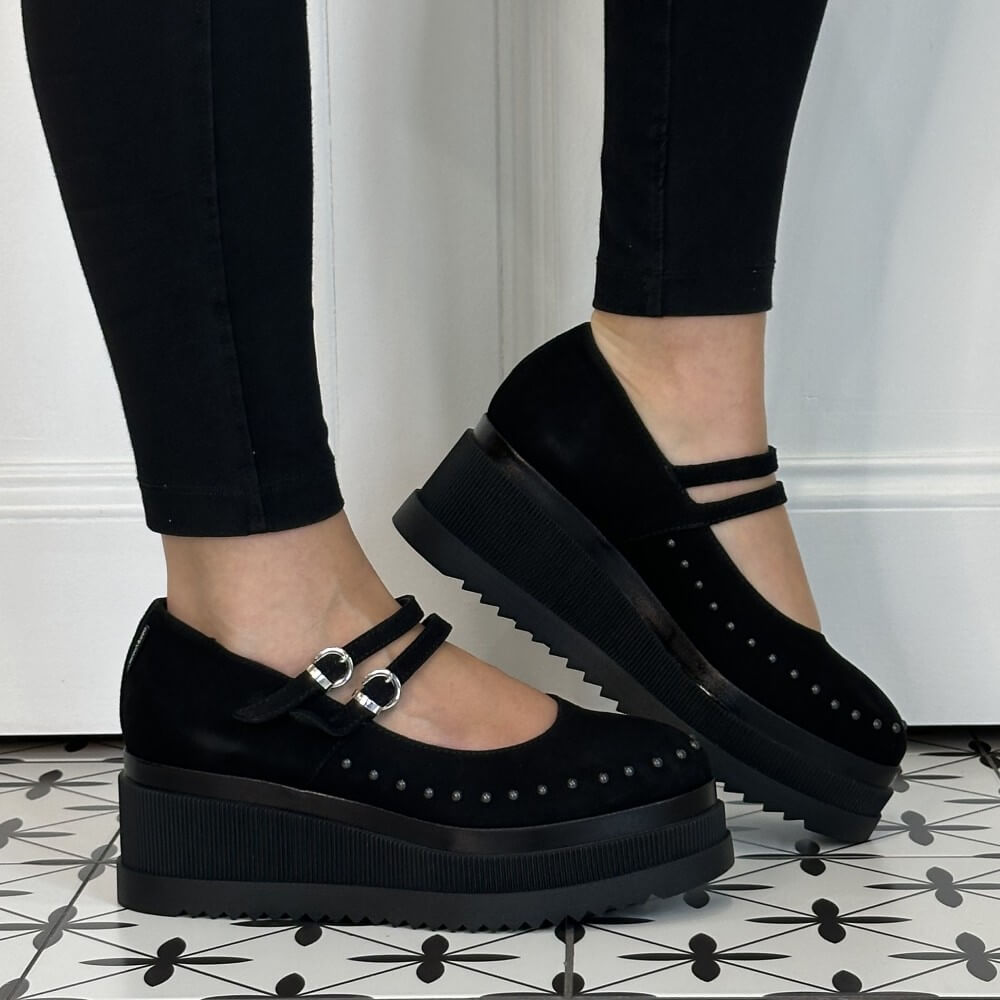 Marco Moreo Black platform Mary Jane shoes with double straps and silver buckles, shown on a model wearing black leggings against a white panelled wall and patterned tile floor.