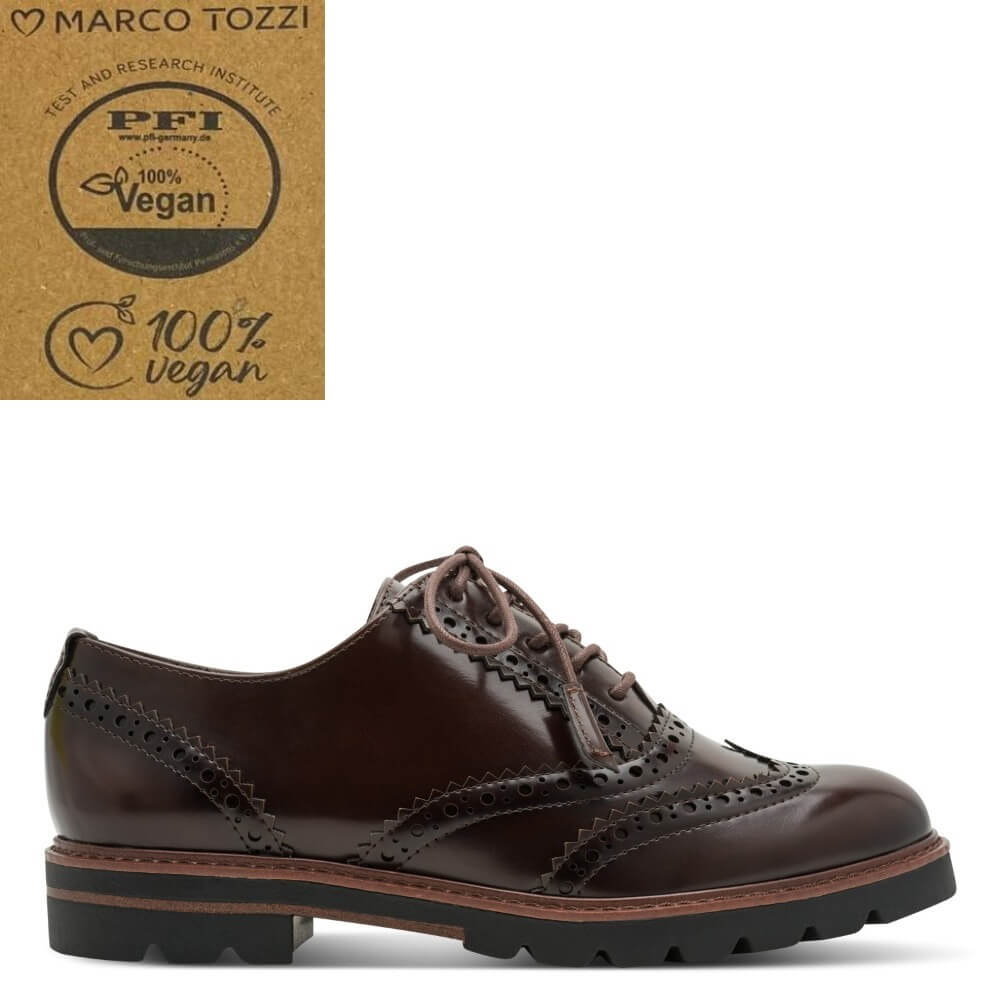 A dark brown leather oxford wingtip shoe with decorative brogue perforations and a chunky black lug sole, shown on a white background. A brown cardboard certification badge for Marco Tozzi's PFI 100% Vegan test is shown in the top-left corner.