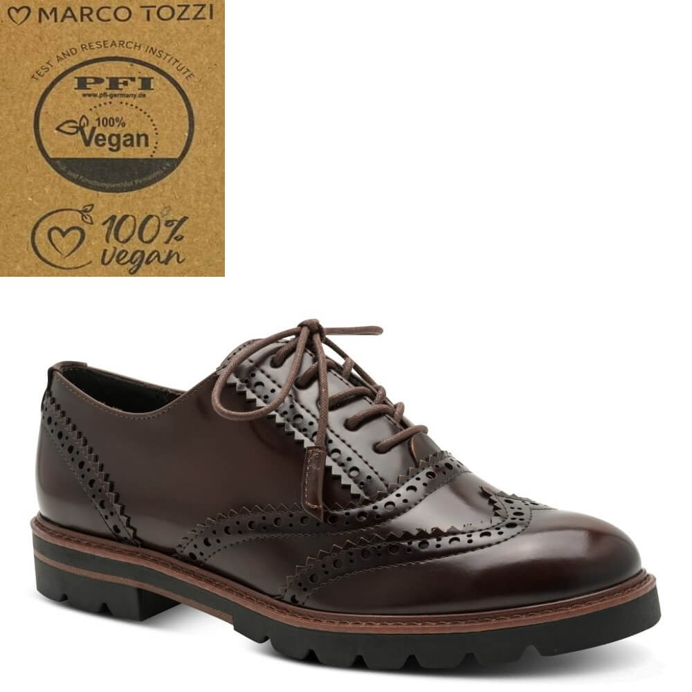 A dark brown leather oxford wingtip shoe with decorative brogue perforations and a chunky black lug sole, shown on a white background. A brown cardboard certification badge for Marco Tozzi's PFI 100% Vegan test is shown in the top-left corner.