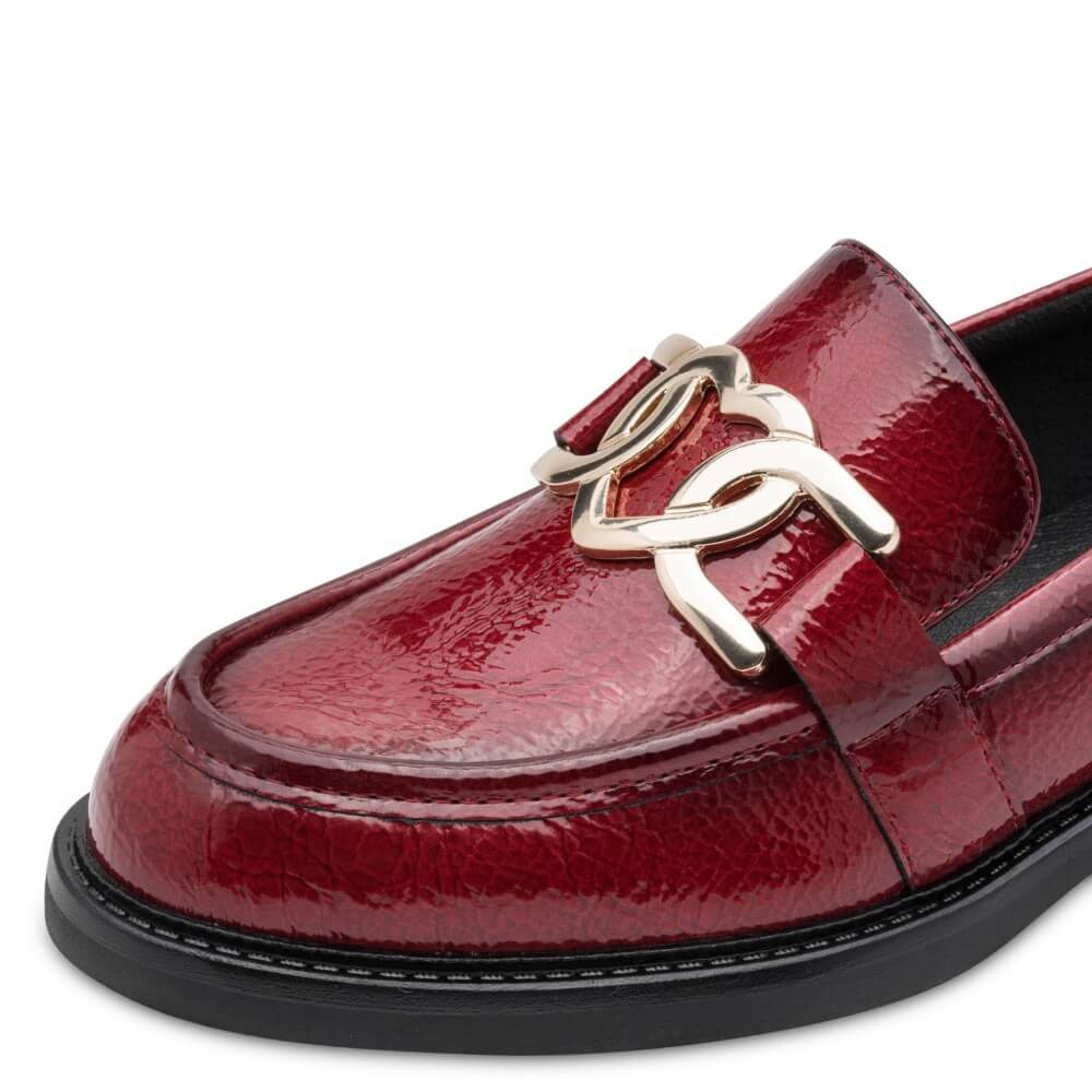 A close-up of a Marco Tozzi red patent leather loafer featuring a prominent metallic gold interlocking rings buckle on the strap, set against a plain white background.