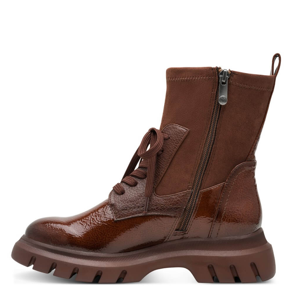 A Marco Tozzi brown ankle boot with a mix of glossy patent Vegan leather and suede panels, featuring side laces, a side zipper, and a chunky lug sole, shown against a plain white background.