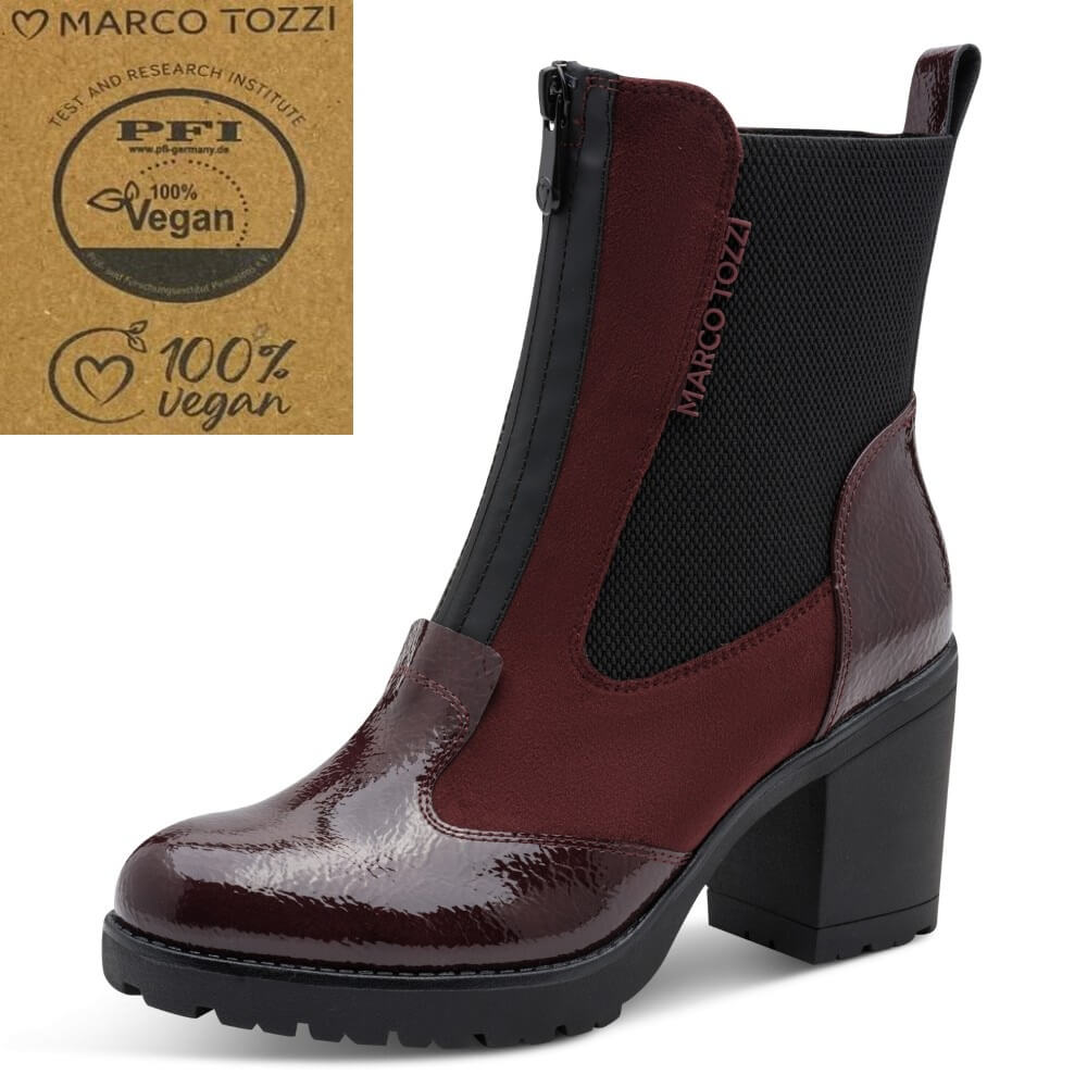 A Marco Tozzi ankle boot with a glossy burgundy and black color-blocked design, featuring a black mesh side panel and a chunky black block heel. A brown cardboard '100% vegan' certification badge is shown in the top-left corner.