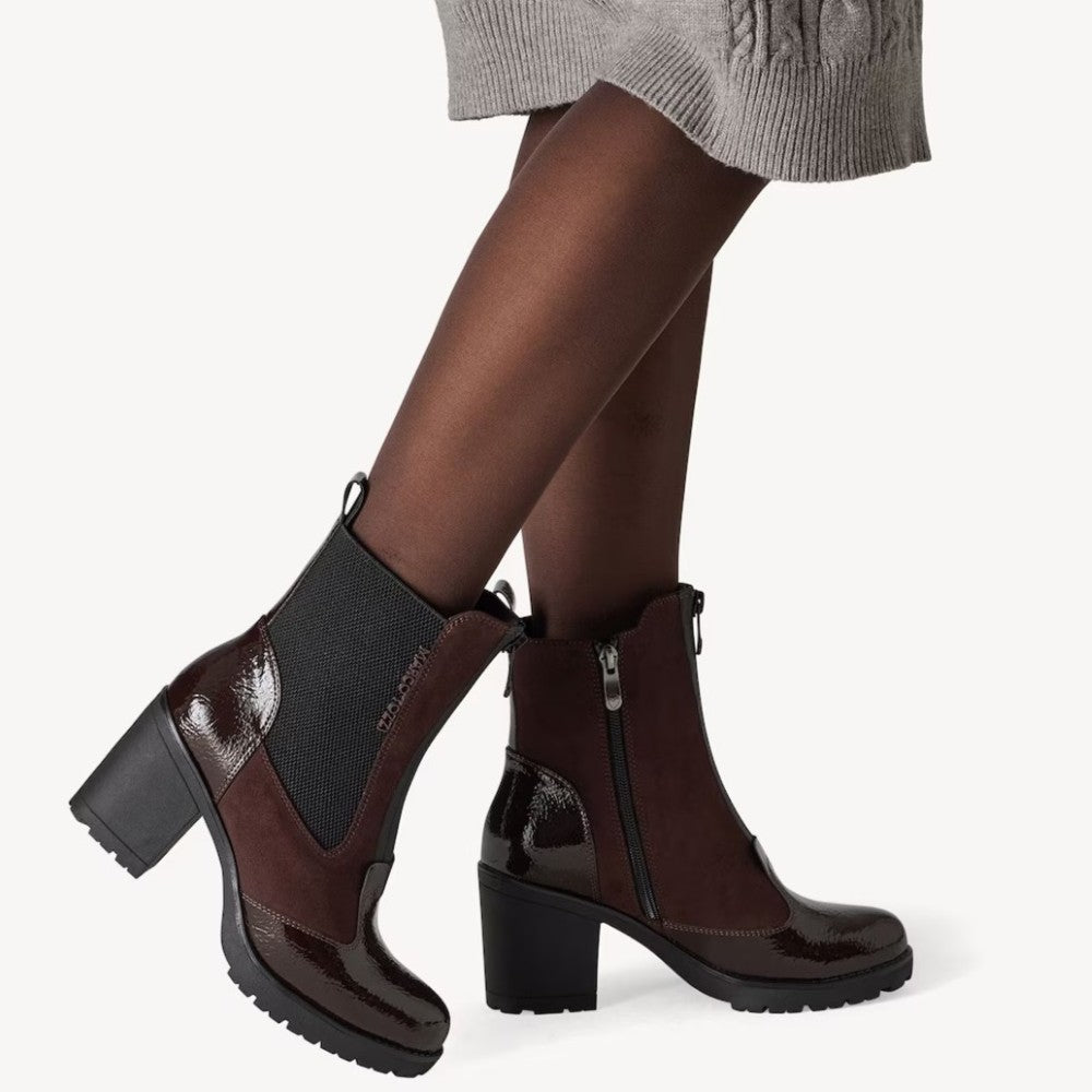 A pair of women's ankle boots in dark brown and black, shown on a model wearing sheer black tights and a gray skirt against a light gray background.