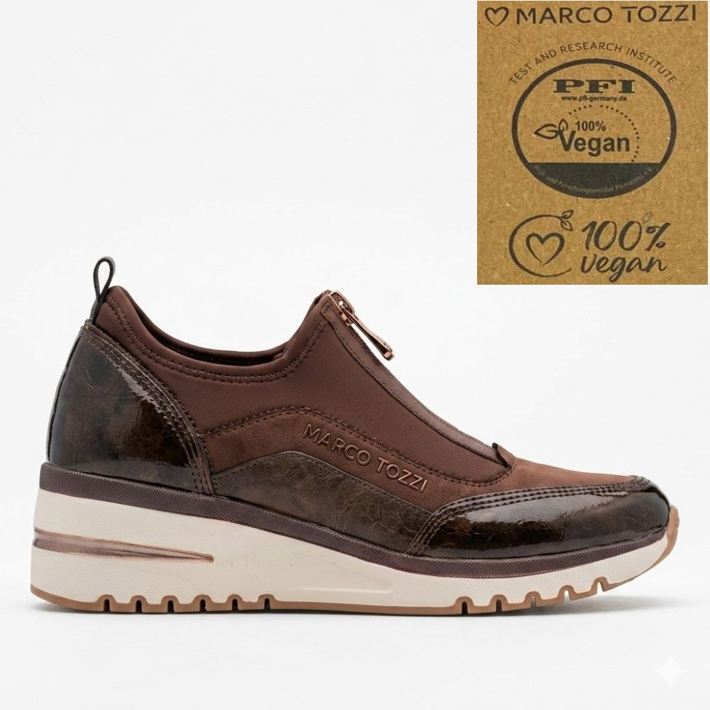 A brown slip-on sneaker by Marco Tozzi, featuring a textured faux-leather upper, a centre zipper, and a thick cream-colored platform sole. A certification badge for '100% Vegan' is shown in the top right corner against a white background.
