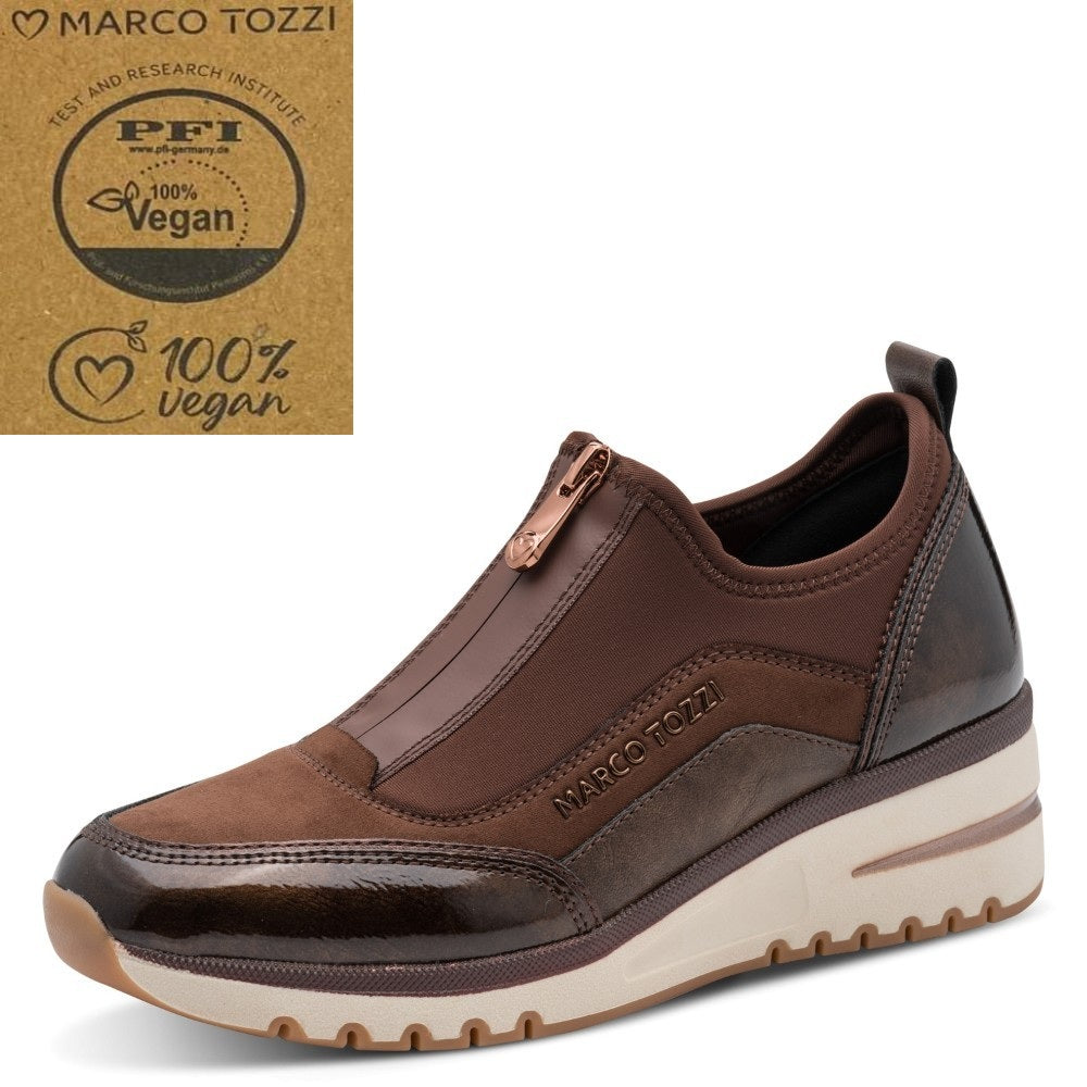 A brown slip-on sneaker by Marco Tozzi, featuring a textured faux-leather upper, a front zipper, and a thick cream-colored platform sole. A certification badge for '100% Vegan' is shown in the top left corner against a white background.