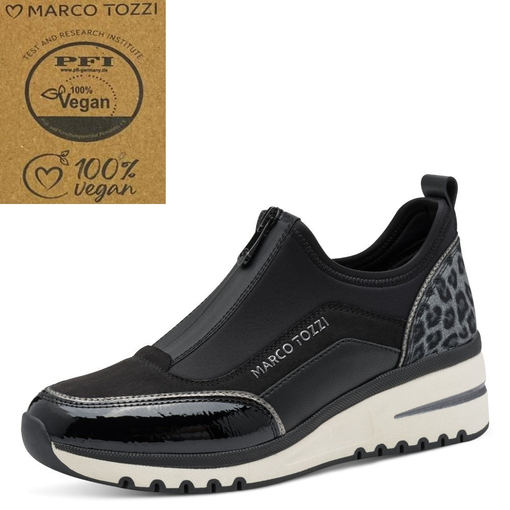 A black slip-on sneaker by Marco Tozzi, featuring a glossy patent leather toe cap, a textured black upper, and a gray leopard print heel panel. The shoe is shown on a white background with a brown cardboard '100% vegan' certification badge in the top-left corner.