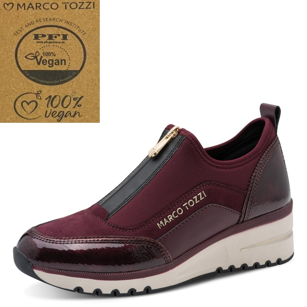 A low-top slip-on sneaker by Marco Tozzi in burgundy and black, featuring a white midsole and a gold zipper. An inset badge on the top right displays a '100% Vegan' certification logo.