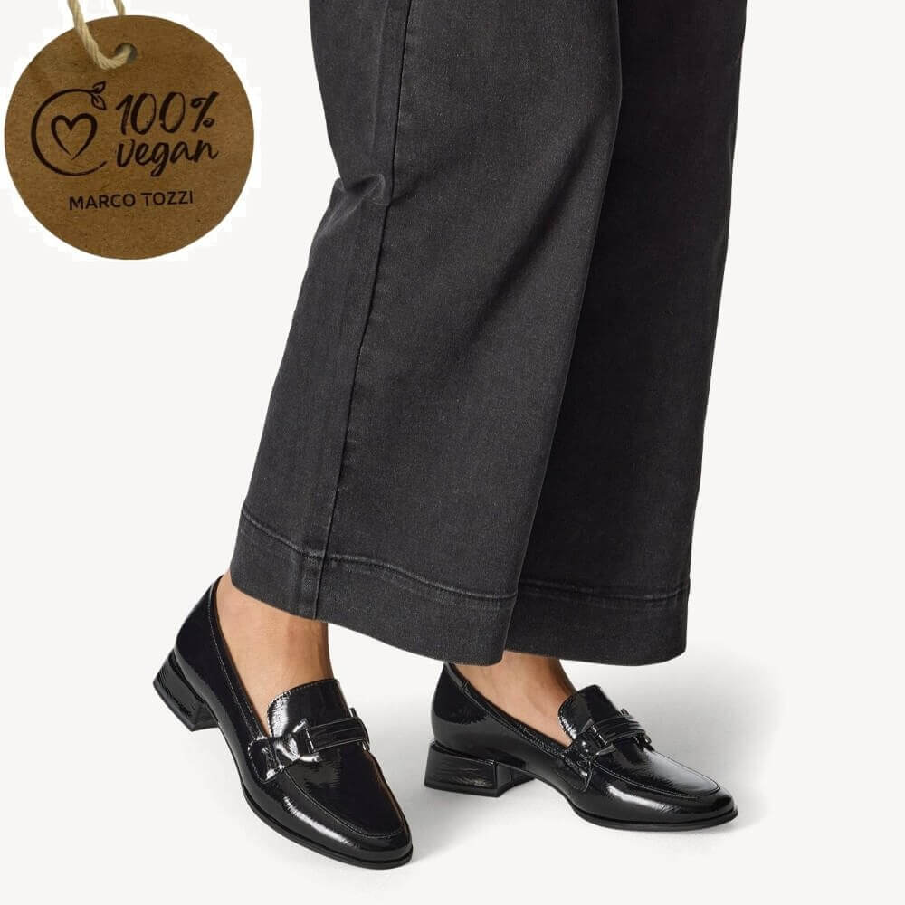 Black loafers worn with dark gray pants, featuring a '100% vegan' tag from Marco Tozzi.
