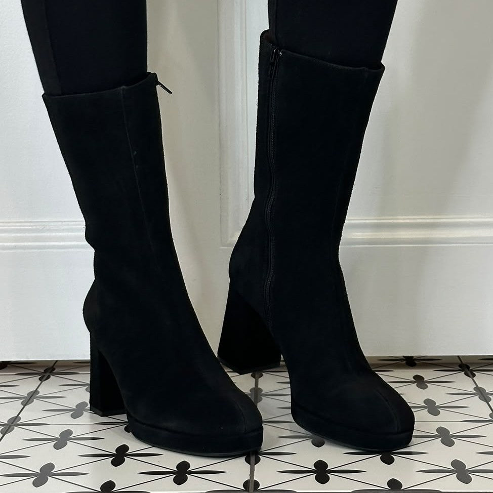 A pair of Marian black suede mid Calf boots with a block heel and side zipper, shown on a person wearing black leggings against a white wall and patterned tile floor.