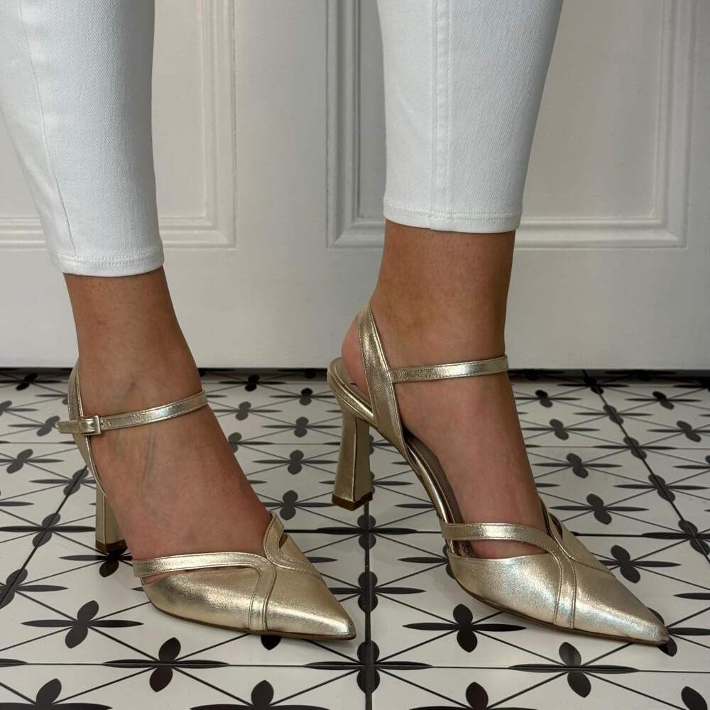 Marian Gold high-heeled shoes worn with white pants on a patterned floor.