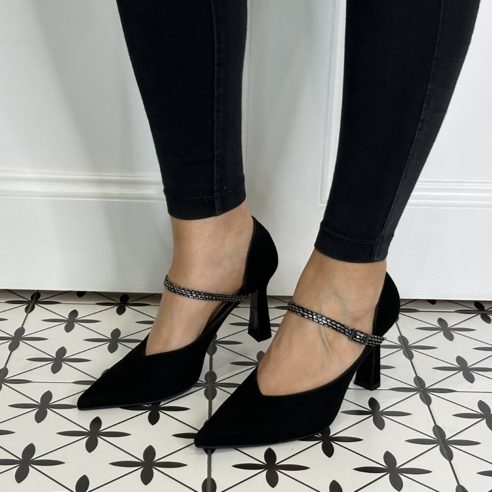 Black suede leather high-heeled shoes with ankle straps worn by a person on a patterned floor.