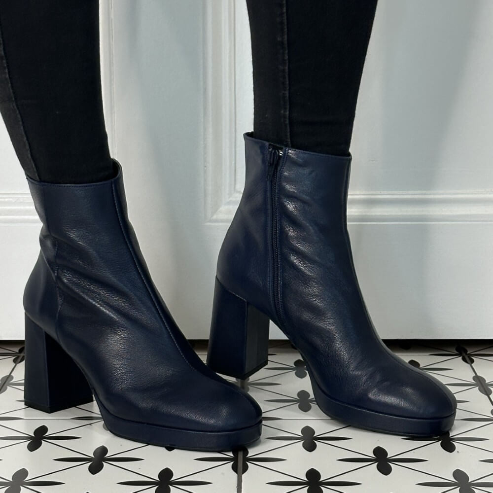 Navy blue leather ankle boots worn with black pants on a patterned floor.