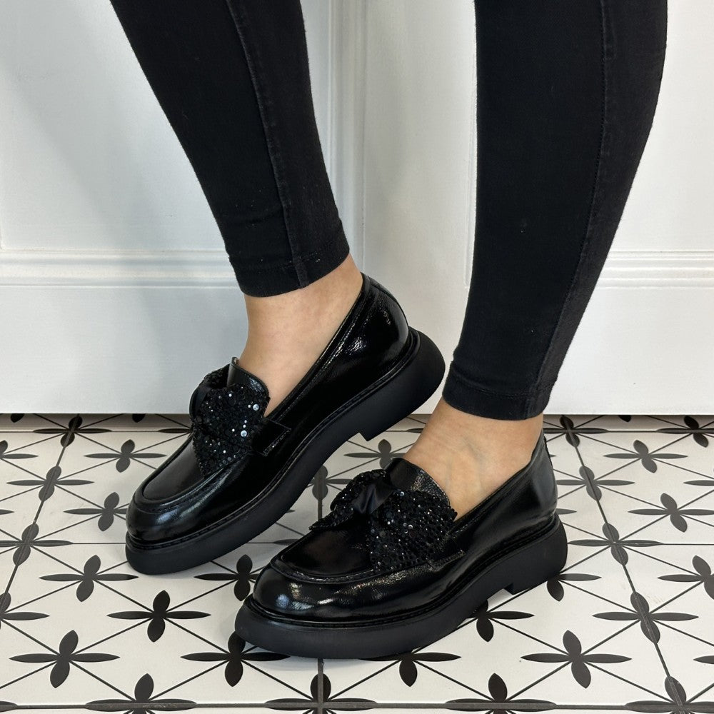 Black loafers with Diamante bow embellishments worn by a person on a patterned floor.