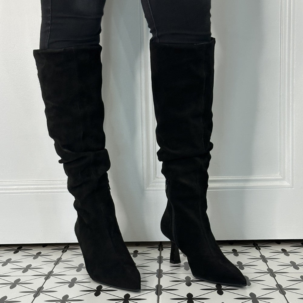 Black suede knee-high boots worn with black pants on a patterned floor.