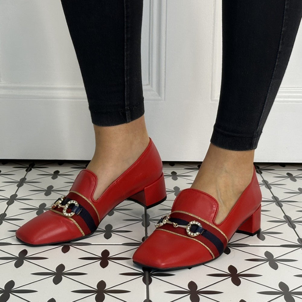 Red leather loafers with black and gold accents worn by a person on a patterned floor.