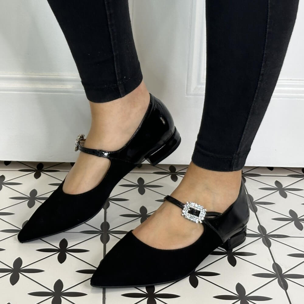 Black pointed-toe flats with crystal buckles worn indoors on a patterned floor.