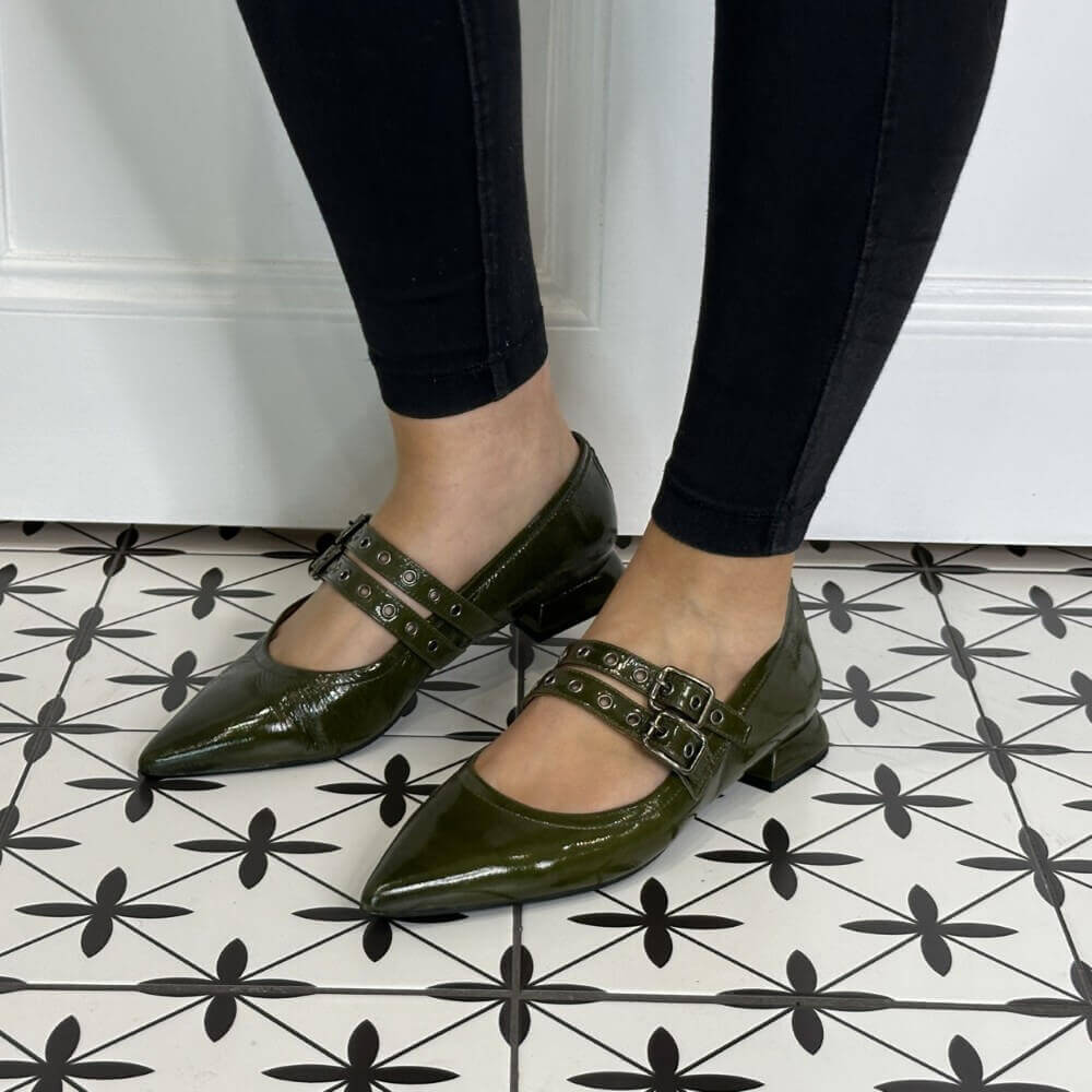 Green patent leather shoes with buckles worn by a person on a black and white patterned floor.