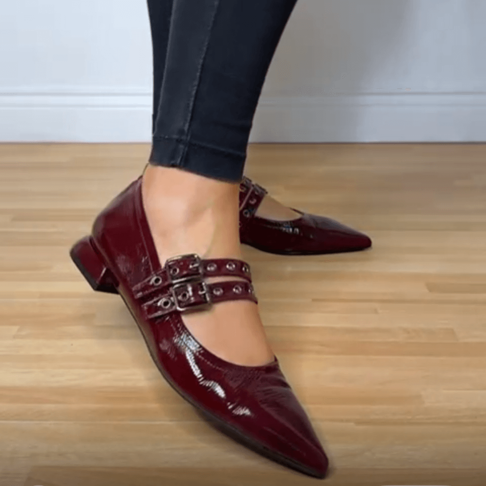 Burgundy pointed-toe shoes with buckles worn by a person on a wooden floor.