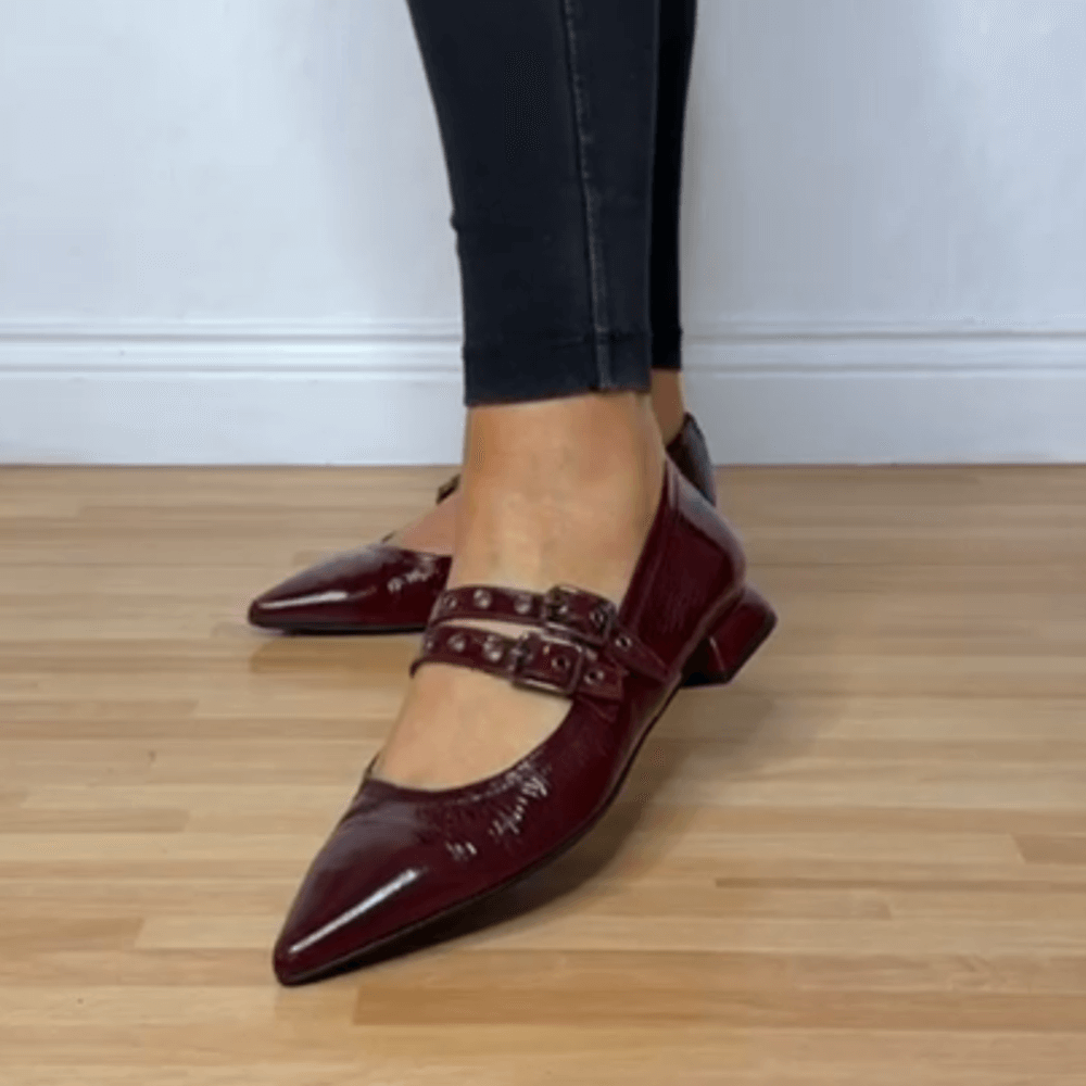 Burgundy pointed-toe shoes with buckles worn by a person on a wooden floor.