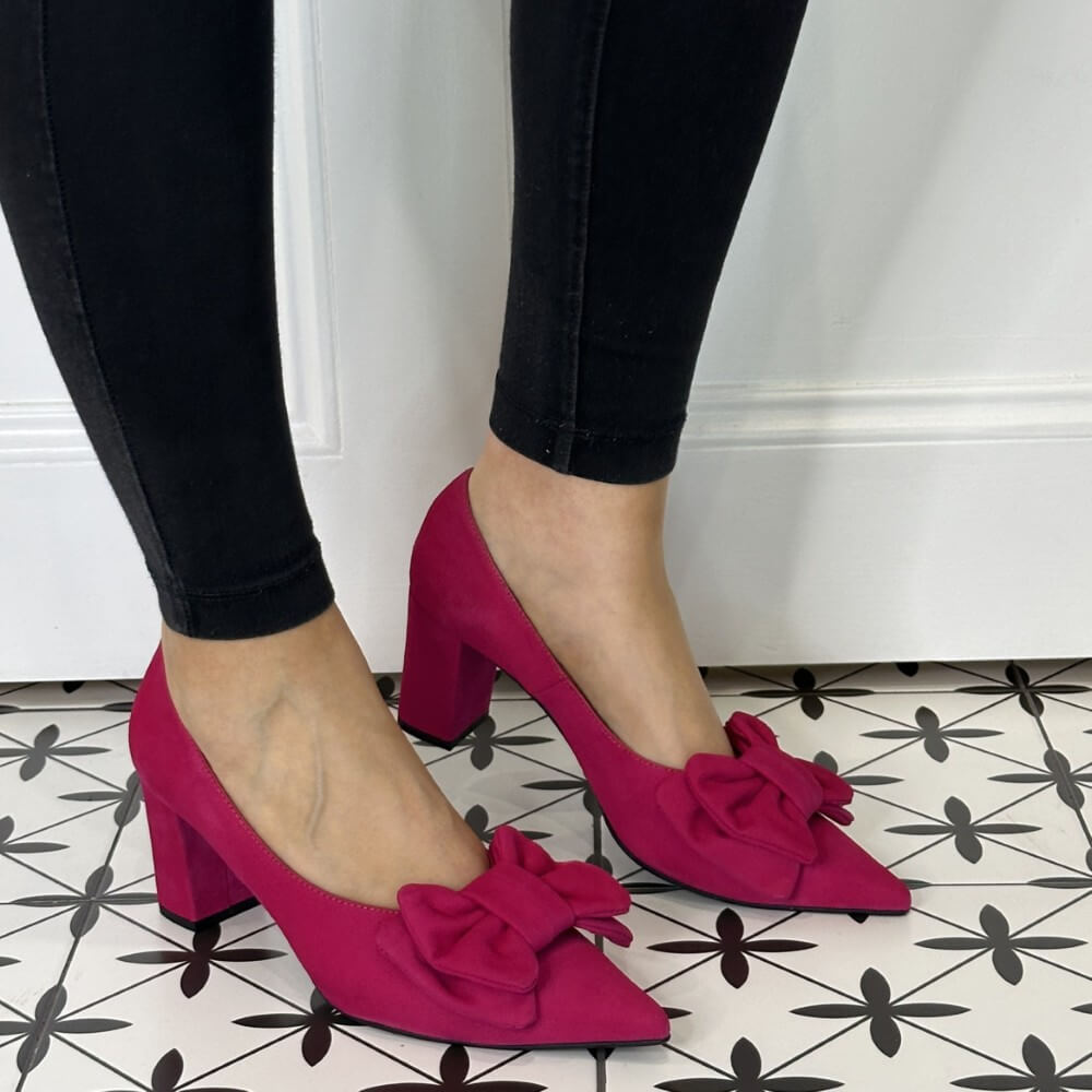 Pink high-heeled shoes with bows worn by a person on a patterned floor.
