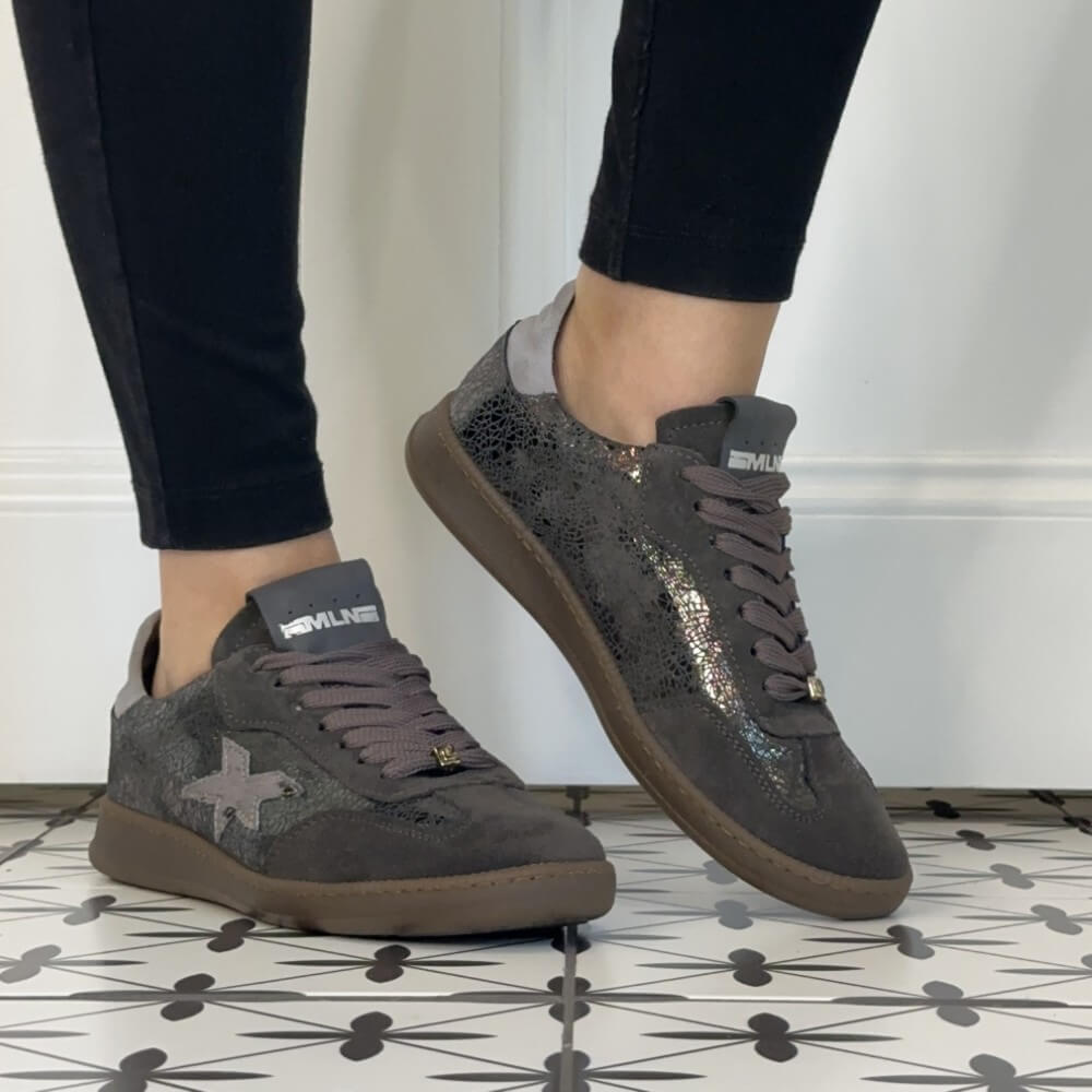 A pair of low-top Meline Retro sneakers with a dark grey glittery upper, brown suede-like overlays, and grey laces, shown on a person wearing black pants standing on a patterned tile floor.