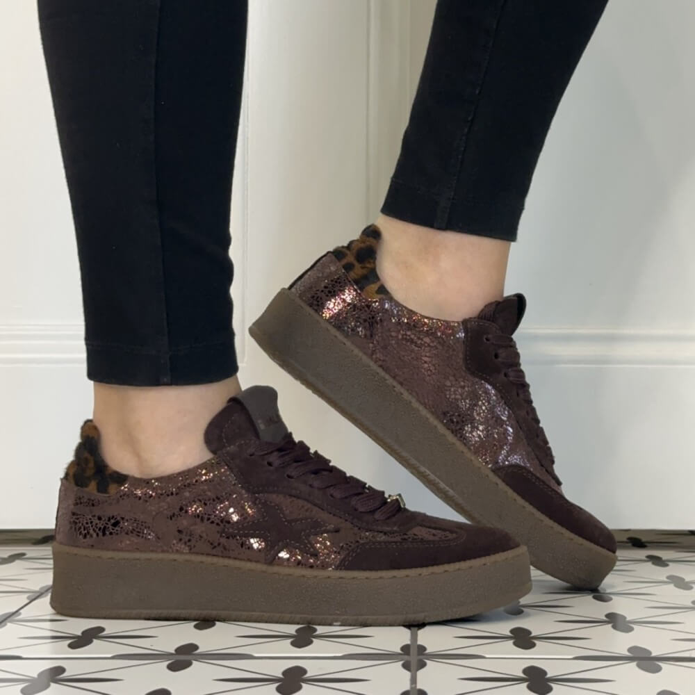 A pair of Meline Retro low-top platform sneakers with dark brown suede and shimmering metallic sequins, featuring a leopard-print heel accent and thick brown soles, shown on a person wearing black pants.