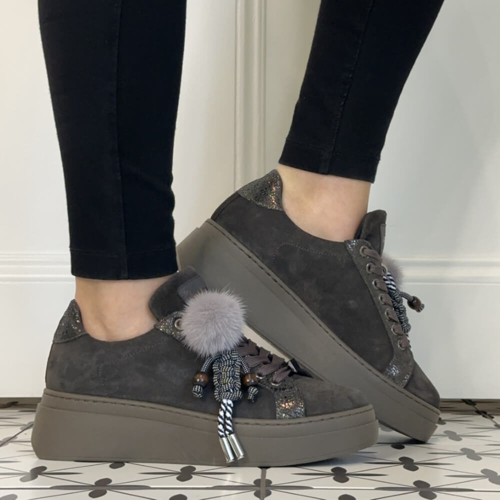 A person wearing Meline Retro dark grey suede platform sneakers with decorative tassels and a fluffy pom-pom, paired with black leggings, standing on a patterned tile floor.