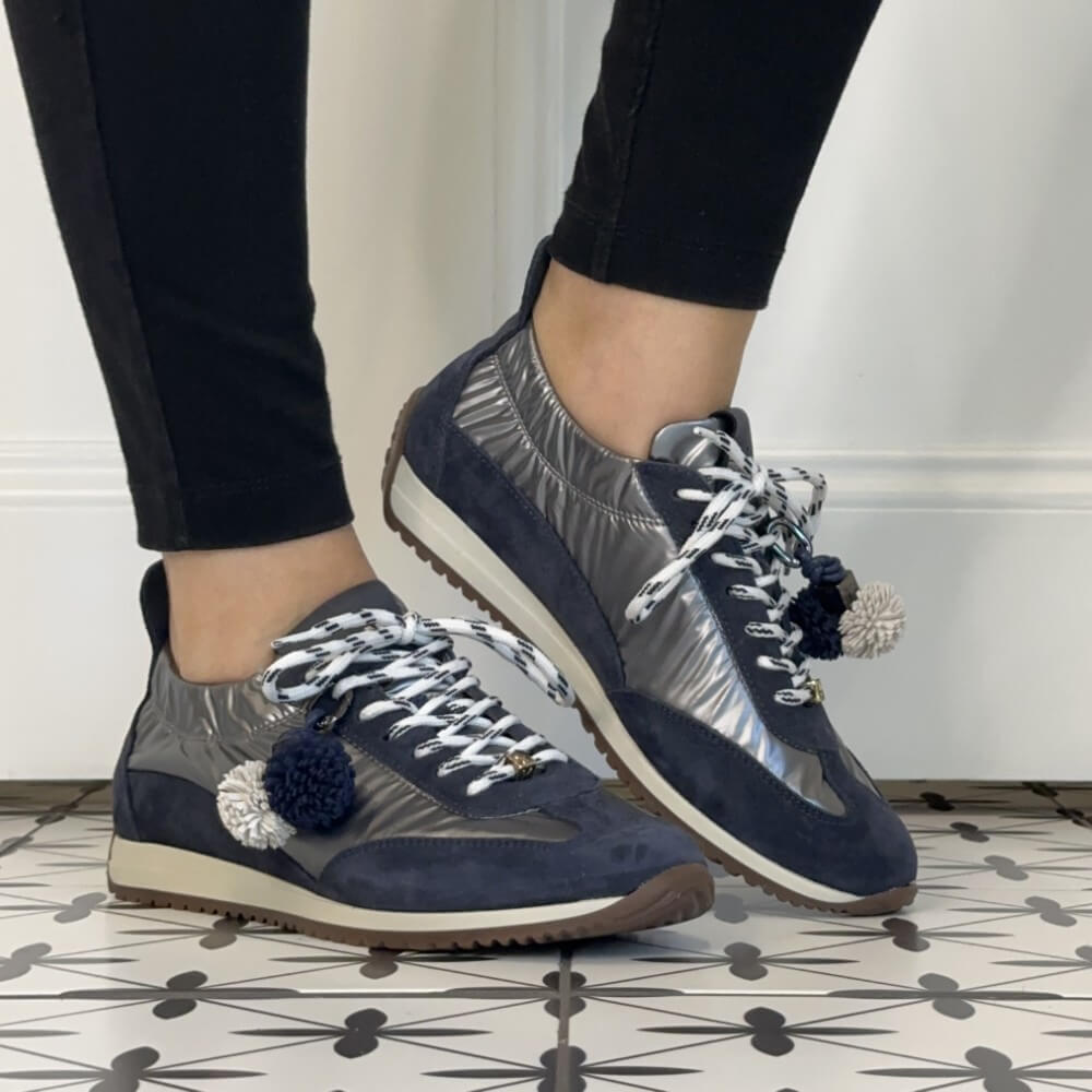 A person wearing Meline Retro navy and metallic silver low-top sneakers with white patterned laces, white pom-pom accents, and black leggings, standing on a black-and-white patterned tile floor.