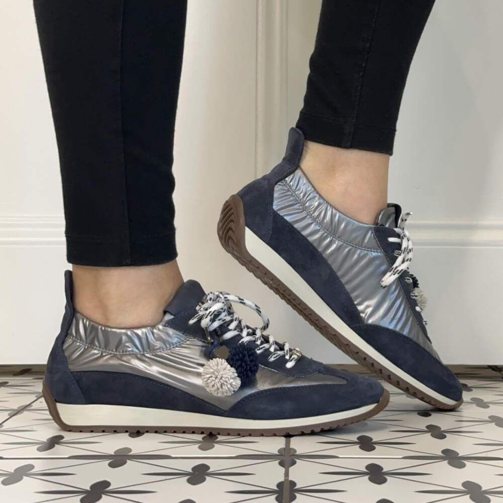 A person wearing Meline Retro navy and metallic silver low-top sneakers with white patterned laces, white pom-pom accents, and black leggings, standing on a black-and-white patterned tile floor.