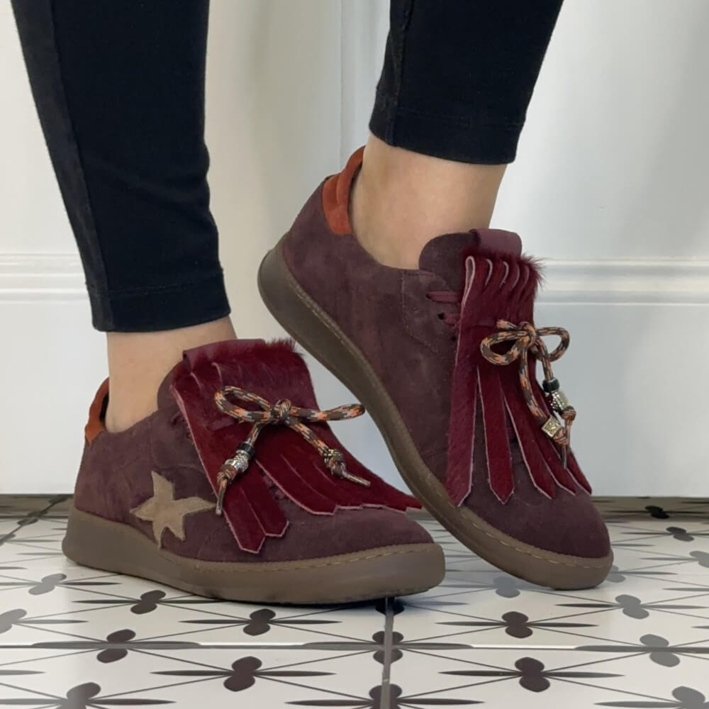 A pair of women's Meline Retro low-top suede sneakers in a deep burgundy colour, shown being worn with black leggings. The shoes feature a beige star on the side, red fringe tassels with beads, and a brown sole.