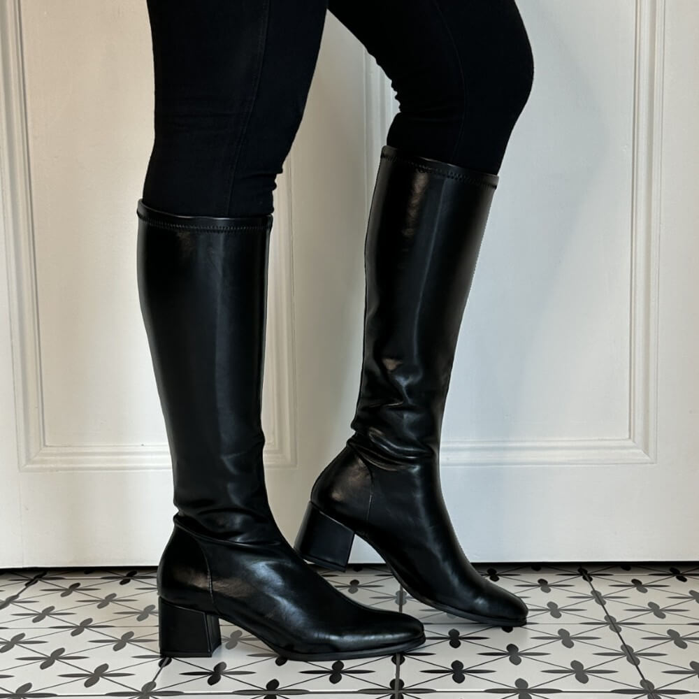 A pair of Miss Elastic black knee-high stretch leather effect boots with block heels, shown on a model wearing black leggings against a white panelled wall and a black-and-white patterned tile floor.