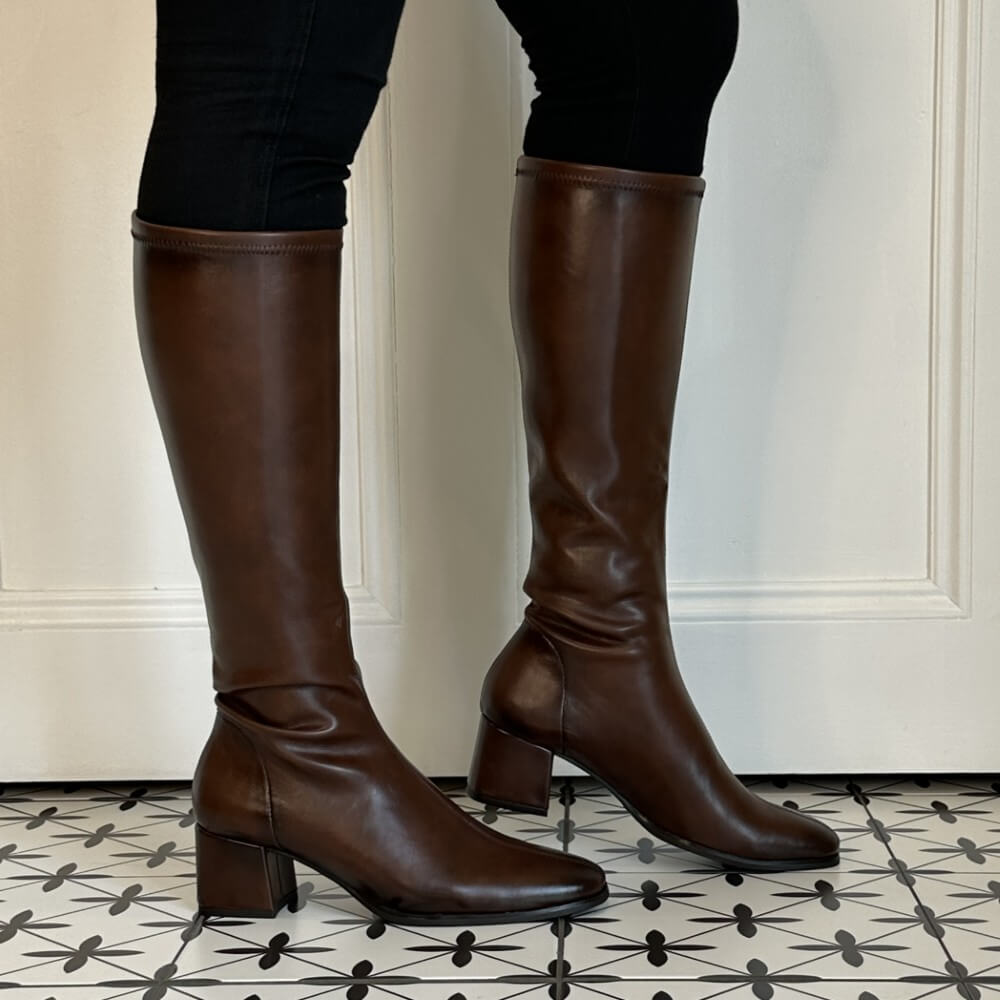 A pair of Miss Elastic dark brown knee-high PU leather boots with block heels, shown on a model wearing black pants against a white panelled door and patterned tile floor.