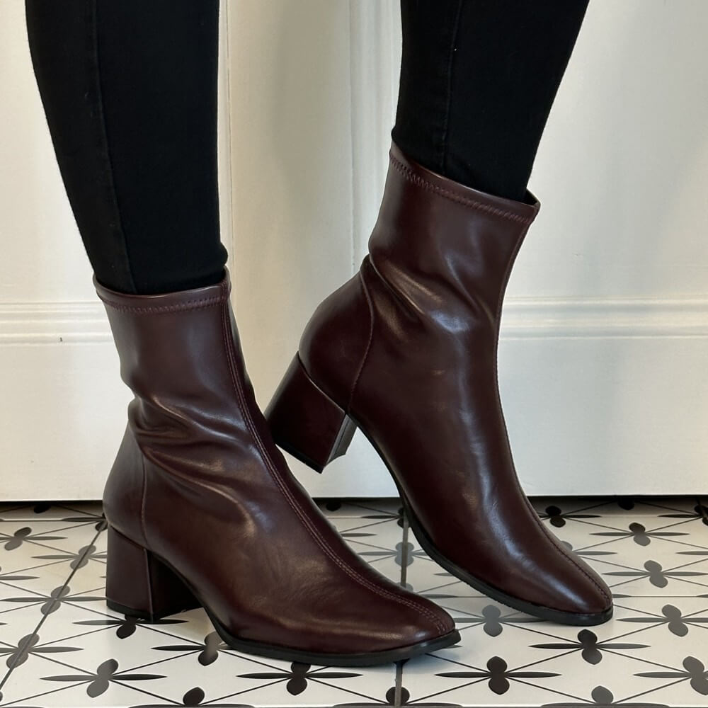A pair of Miss Elastic Wine ankle boots with block heels, worn with black leggings on a patterned black-and-white tile floor in front of a white wall.