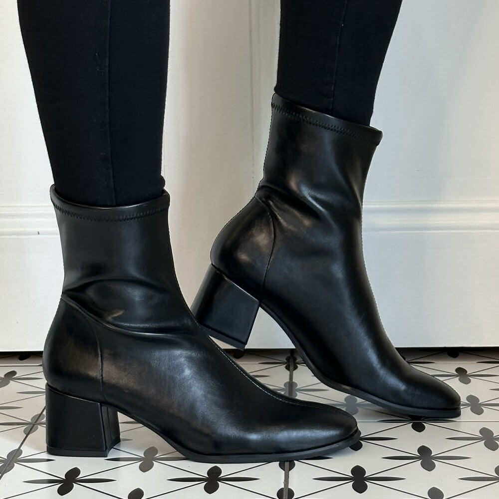 A pair of Miss Elastic black ankle boots with block heels, shown on a model wearing black leggings. The boots are positioned on a black and white patterned tile floor against a white wall.