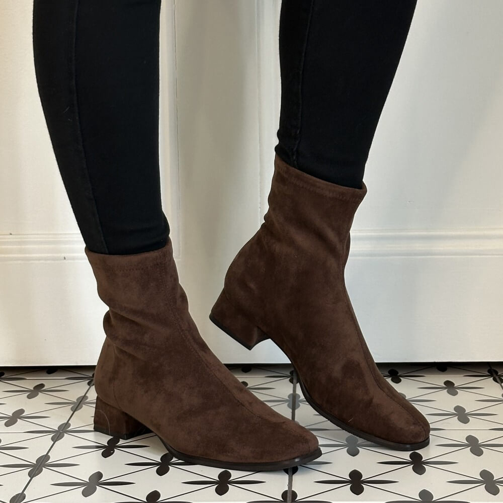 A pair of Miss Elastic dark brown suede ankle boots with low block heels, shown on a model wearing black leggings standing on a black and white patterned tile floor.