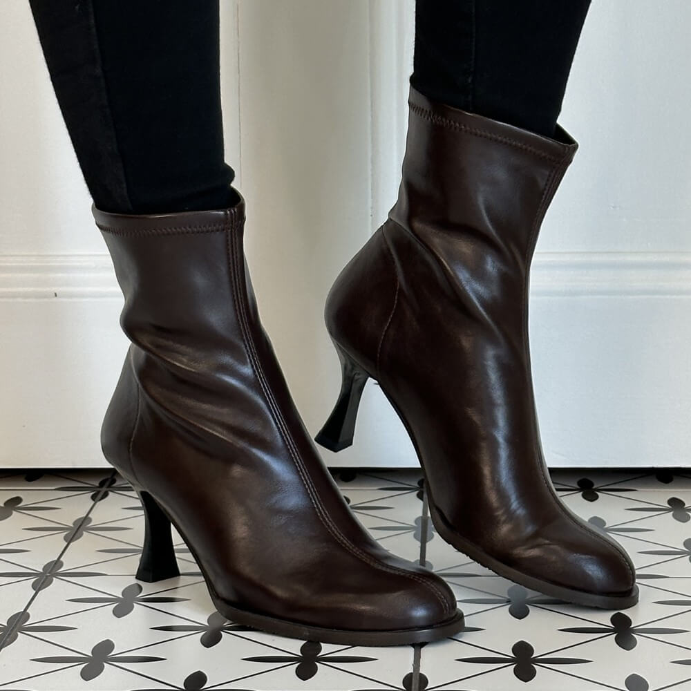 A pair of Miss Elastic dark brown stretch ankle boots with kitten heels, shown on a model wearing black pants. The boots are positioned on a black and white patterned tile floor against a white wall.