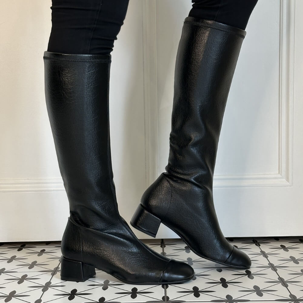 A pair of Miss Elastic black leather knee-high boots with low block heels, shown on a model wearing black leggings against a white panelled door and a black-and-white patterned tile floor.