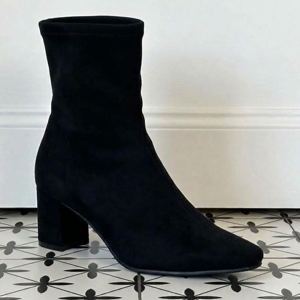 A Miss Elastic black stretch suede effect ankle boot with a chunky block heel and pointed toe, photographed on a patterned black-and-white tile floor against a white wall.