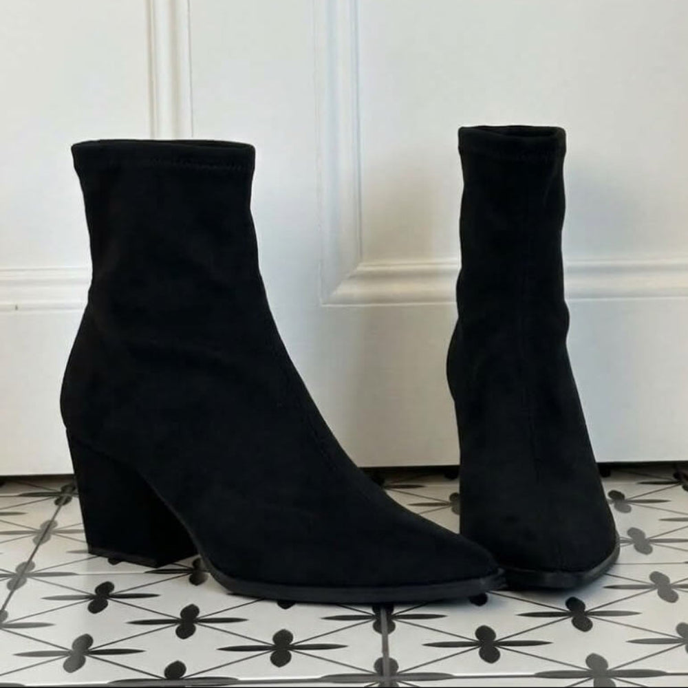 A pair of Miss Elastic black stretch ankle boots with block heels, displayed on a black and white patterned tile floor in front of a white panelled wall.